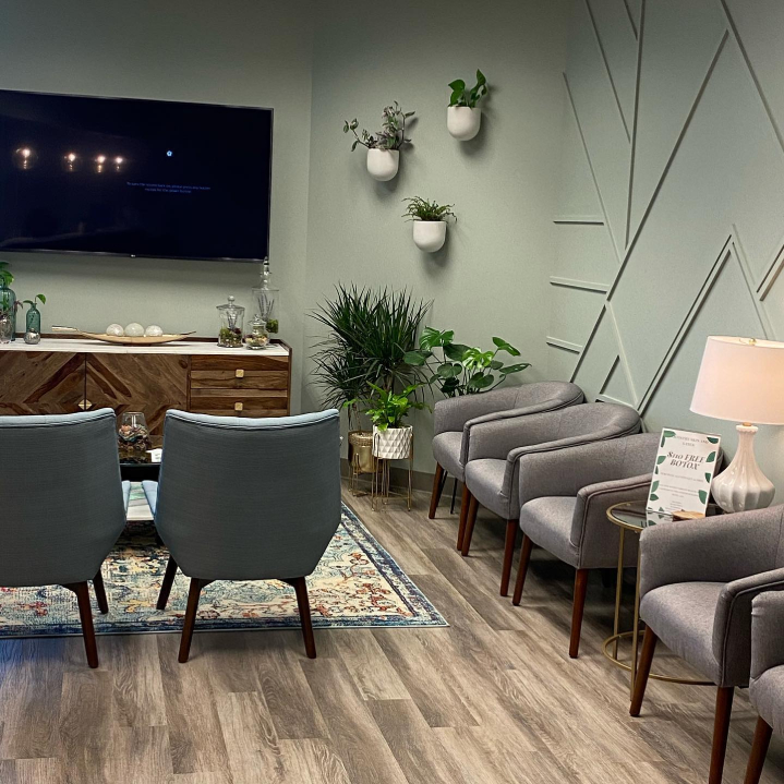 An inviting and cozy patient waiting area with plants, a tv, comfortable seating, and lovely green walls at Artistry Skin & Laser.